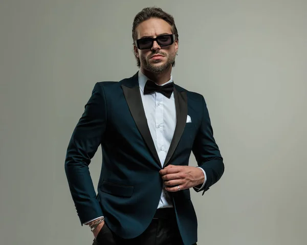 elegant young man in tuxedo with sunglasses holding hand in pocket and unbuttoning tux in front of grey background