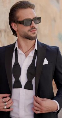 Kendine güvenen papyonlu bir moda adamı takım elbisesini kapatmaya ve ayarlamaya çalışıyor, güneş gözlüğünü tamir ediyor, aşağı bakıyor ve dışarı çıkıyor.