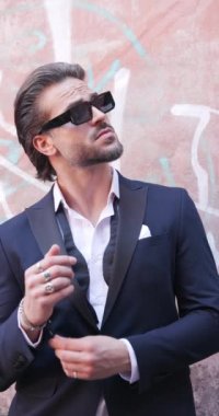 Meraklı, güneş gözlüklü bir iş adamı eski şehre bakıyor, takım elbisesini ayarlıyor ve elini cebine atarak yürüyor.