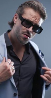 Güneş gözlüklü çekici bir adam takım elbisesini iliklerken yana bakıyor, gri arka planda el ele tutuşuyor ve el ele tutuşuyor.