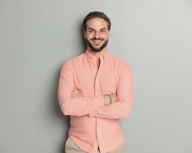 Ekose gömlekli gururlu bir gencin portresi kollarını kavuşturuyor, gri arka planda mutlu ve gülümsüyor.