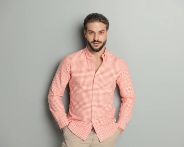 Ekose gömlekli seksi sakallı adam ceplerinde el ele tutuşuyor ve gri arka plan önünde poz veriyor.