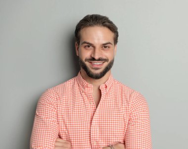 Mutlu sıradan bir adamın portresi sakallı kollarını kavuşturup stüdyoda gri arka planda gülümsüyor.