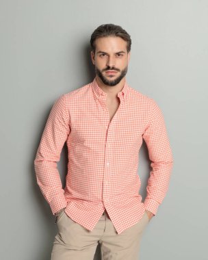 Yakışıklı, sıradan, sakallı, el ele tutuşan ve gri arka planda ileriye bakan bir adam.