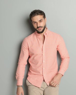 Tıraşsız, kendine güvenen, gömlek giyen ve stüdyoda gri arka planda poz veren genç bir adamın portresi.