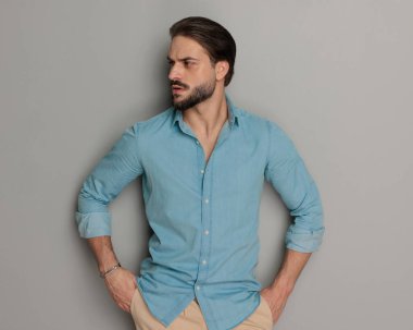 Sakallı yakışıklı genç adam gri arka planda el ele tutuşup yan tarafa bakıyor. 