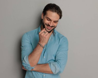 Heyecanlı sıradan bir adam kollarını kavuşturuyor, aşağı bakıyor ve gri arka planın önünde gülüyor. 