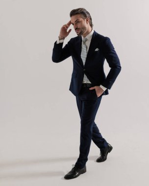 Üst sınıf genç adam tapınağa dokunuyor, yan tarafa bakıyor ve stüdyoda gri arka planda el ele yürüyor.