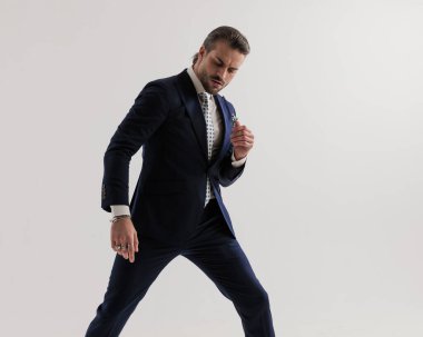 Kravat ve mendille donanma mavisi takım elbiseli seksi havalı iş adamı aşağı bakıyor ve gri arka planda poz verirken kollarını tutuyor.