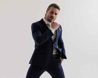 Seksi, zarif bir adam, çenesine dokunuyor ve elini cebine atıyor ve gri arka planda poz veriyor. 