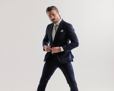 Gözlüklü zarif seksi adam gri arka planda üst sınıf takımını iliklerken havalı poz veriyor.