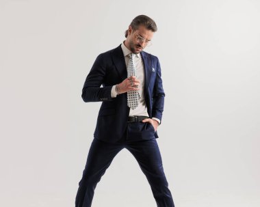 Gri arka planda kravatını düzeltirken el ele tutuşup aşağı bakan gözlüklü seksi, üst sınıf bir adam. 