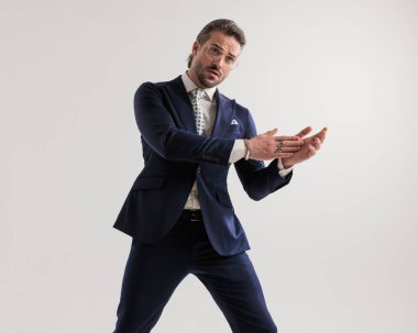 Avuçlarını ovalarken ve gri arka planda kendine güvenen bir şekilde poz verirken gözlüklü zarif bir adam.