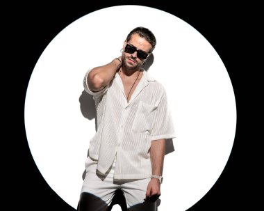 Güneş gözlüklü şık bir adam, boynunun arkasında el ele tutuşuyor ve beyaz arka planda sahne ışıkları önünde kendine güvenen bir şekilde poz veriyor.