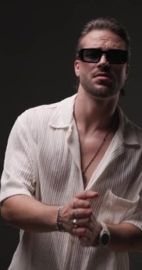 Güneş gözlüklü, çene ve avuçlarına dokunan, düşünceli, yana bakan ve özgüvenle gri arka planda poz veren seksi bir adamın dikey videosu.
