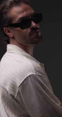 Güneş gözlüklü, gülümseyen, omuzlarına dokunan ve gri arka plandaki çenesine dokunarak dönen sıradan bir gencin arka planı.