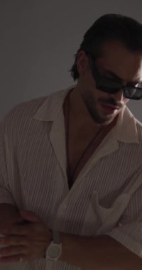Güneş gözlüklü, hareketli, avuçlarını ovuşturan, poz verirken kendine güvenen ve gri arka plana bakan seksi moda adamının dikey videosu.