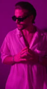 Güneş gözlüklü, el ele tutuşan, güneş gözlüğünü ayarlayan ve gri arka planda renkli ışıkları olan seksi genç adamın dikey videosu.