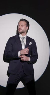 Seksi sakallı adam kollarını ve takım elbisesini düzeltiyor. Düğmelerini açıyor, çenesine dokunuyor ve gri arkaplan önünde düşünüyor.