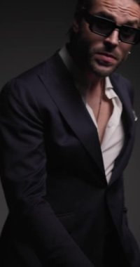 dinamik zarif adam aşağı bakıyor, hareket ediyor, takım elbise ayarlıyor ve kendine güveniyor, dönüyor ve gri arka plana özgüvenle bakıyor.