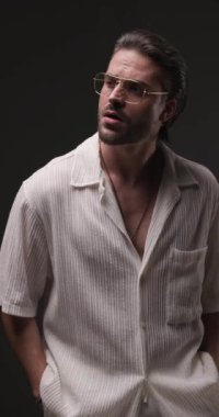 Altın çerçeveli gözlüklü, el ele tutuşan, aşağıya bakan, gömleği ayarlayan ve gri arka planda hareket eden seksi bir adamın dikey videosu.