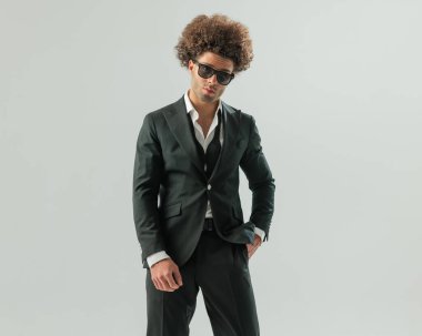 Uzun kıvırcık saçlı, cebinde siyah takım elbiseli, yakalı ve gri arka planda güneş gözlüklü çekici bir adam.