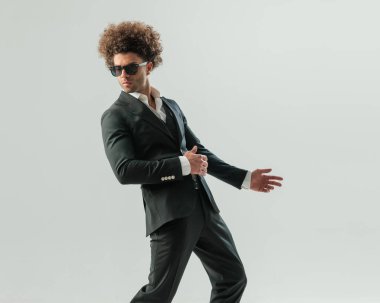 Güneş gözlüklü çekici bir adam omuzlarının üzerinden bakıyor ve kollarını kaldırıp gri arka planda poz veriyor.