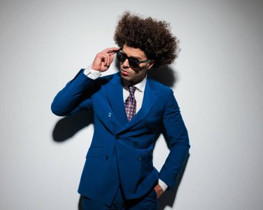 Uzun kıvırcık saçlı, cebinde el ele tutuşan ve gri arka plana bakarken güneş gözlüğü takan zarif genç iş adamının portresi. 