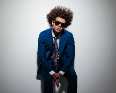 Mavi takım elbiseli zarif bir adam avuçlarını bir arada tutuyor, duvara yaslanıyor ve gri arka planda serinliyor. 