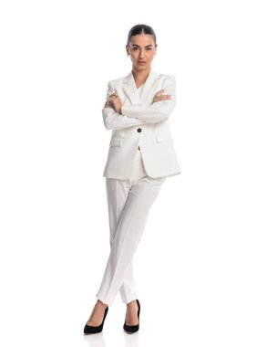 Beyaz takım elbiseli kendine güvenen genç bir kadın stüdyoda beyaz arka planda dikilirken kollarını kavuşturup ileriye bakıyor.