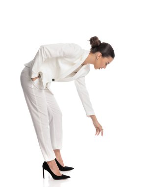 Beyaz takım elbiseli zarif bir kadının el ele tutuşup, beyaz arka planda yerden bir şey almak için eğilmesinin yan görüntüsü.