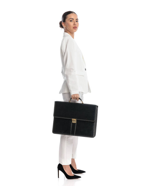 side view of confident young woman in white suit with bun hair holding suitcase and posing in front of white background 