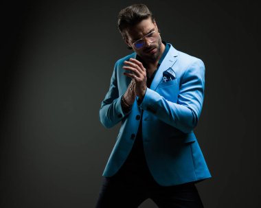 Mendilli, gözlüklü, koyu gri arka planda avuçlarını ovuşturan mavi takım elbiseli üst sınıf bir iş adamı.