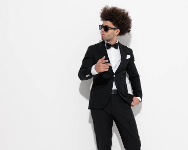 Güneş gözlüklü zarif genç işadamı bir elini cebine sokup diğeriyle gri arka planda el kol geziyor.