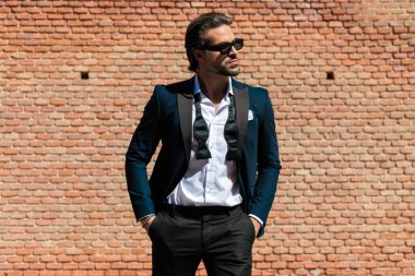 Takım elbiseli, açık yakalı, güneş gözlüklü, cebinde el ele tutuşan şık bir adam.