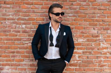 Yakası açık ve güneş gözlüklü havalı moda işadamı eski bir şehirde ceplerinde ellerle poz veriyor.