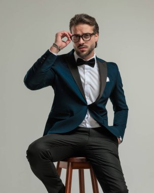 Smokinli çekici, zarif bir adam. El ele tutuşup koyu gri arka planda otururken gözlük takıyor.