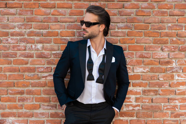 sexy cool man with open collar shirt and sunglasses holding hands in pockets and looking to side in front of stones wall outdoor in an old city