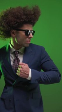 Güneş gözlüklü, takım elbiseli ve kravatlı seksi, üst sınıf bir adam. Renkli arka planda kamera kapalıyken ileriye bakıyor.