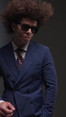 Kibirli, zarif bir adam, el ele tutuşuyor, güneş gözlüğü ve çeneye dokunuyor, kameralar gri arkaplanı kapatırken kendinden emin bir şekilde ileriye bakıyor.