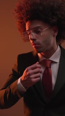 Gözlüklü seksi ve zarif bir adam takım elbisesini ayarlamaya çalışıyor, çenesine dokunuyor ve alevlerin önüne bakarken gözlüklerine bakıyor.