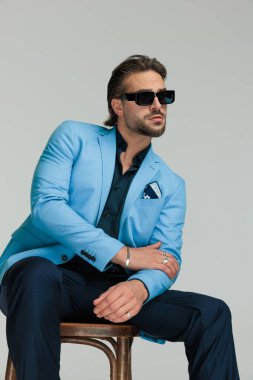 Mavi gömlekli, yakalı, güneş gözlüklü, ahşap koltukta oturan zarif bir moda adamı. Gri arka planda kollarına dokunmaya çalışıyor.