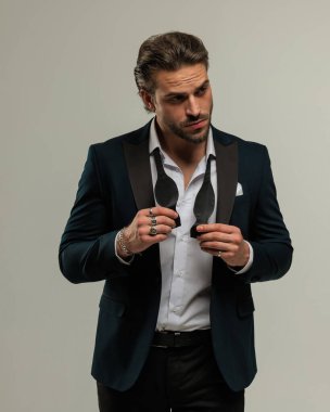 Zarif takım elbiseli yakışıklı genç adam gri arka planda poz verirken papyon takmaya ve düzeltmeye çalışıyor.