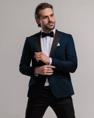 Smokinli kendine güvenen genç işadamı gri arka planda poz verirken kollarını düzeltiyor.