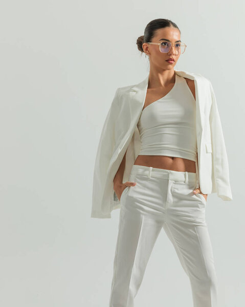 confident smart casual lady with glasses looking away and holding hands in pockets in front of grey background