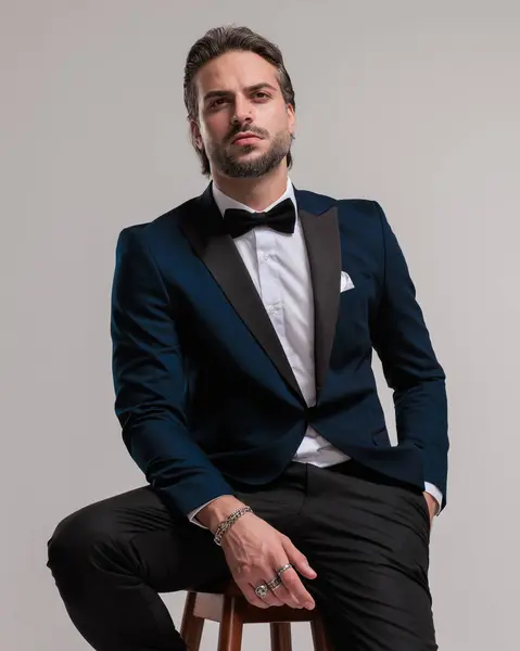 confident high class best man looking up and holding hand in pocket while sitting in front of grey background