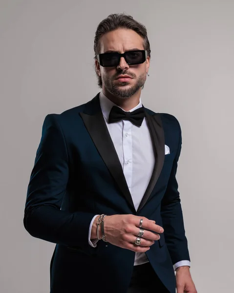 stock image cool fashion man with sunglasses wearing tuxedo and looking forward while posing in a confident way on grey background