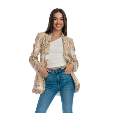 Kendine güvenen, ceketli, kot pantolonunun cebinde el ele tutuşan, ileriye bakan ve beyaz arka planda gülümseyen bir kız.