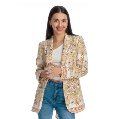 Güzel genç bir kadın öne bakıyor ve gülümsüyor. Poz veriyor ve beyaz arka planın önünde el ele tutuşuyor. 