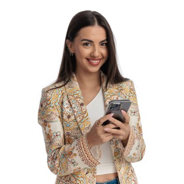 Önüne bakan ceketli mutlu genç kız, elinde telefon, mesajlar, gülümsüyor ve beyaz arka planda poz veriyor.